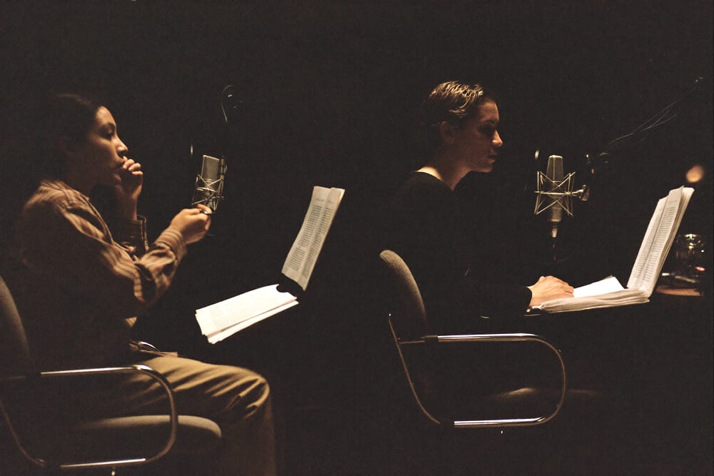 Two people mid-conversation at microphones in a warm, dark recording room.
