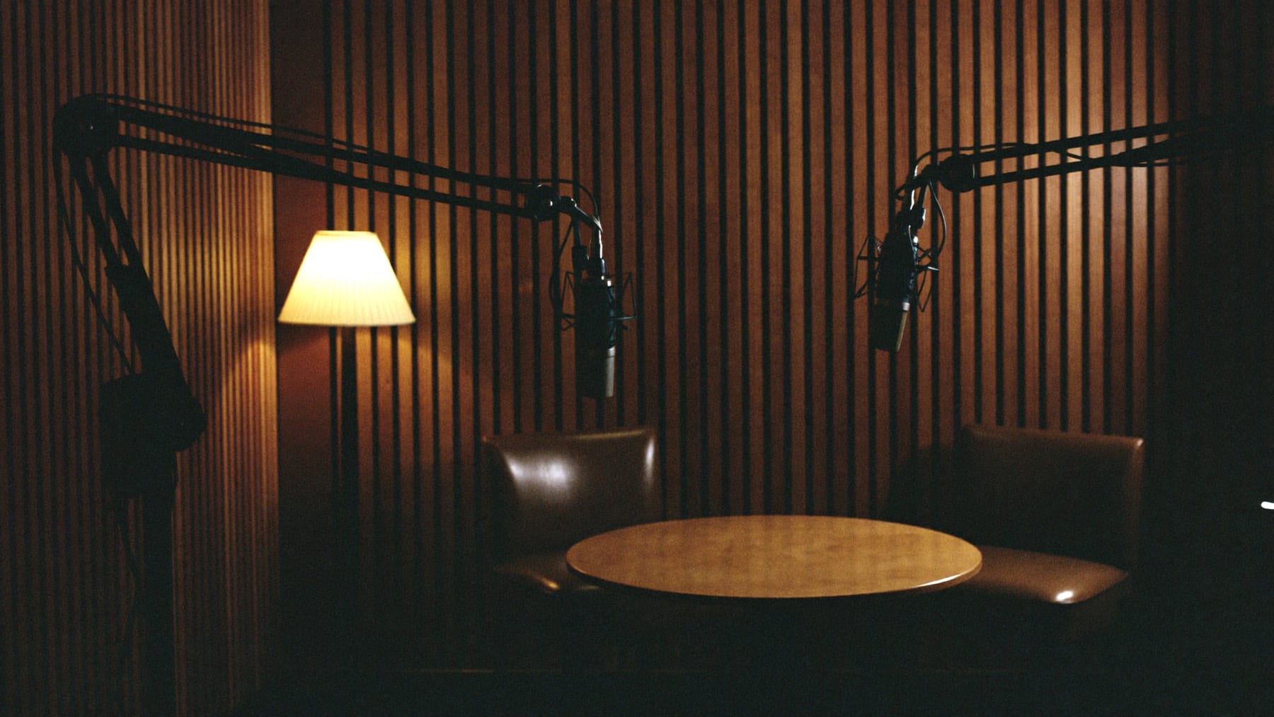 A warm, dimly lit podcast room: two microphones on boom arms over a round table, two empty chairs, slatted wood walls.