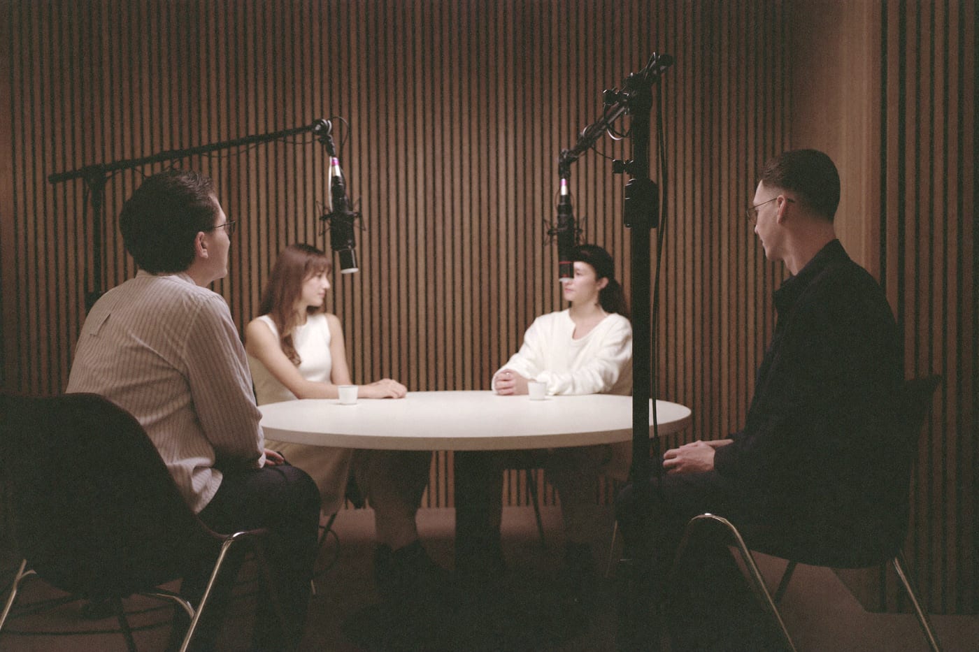 A podcast studio set for four: four microphones on boom arms around a round table, four empty chairs.